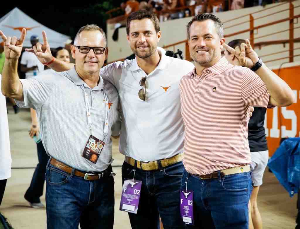David Pierce, Justin McMinn, and Jason McMinn smile and hold up longhorn hook em horns signs at a football game.